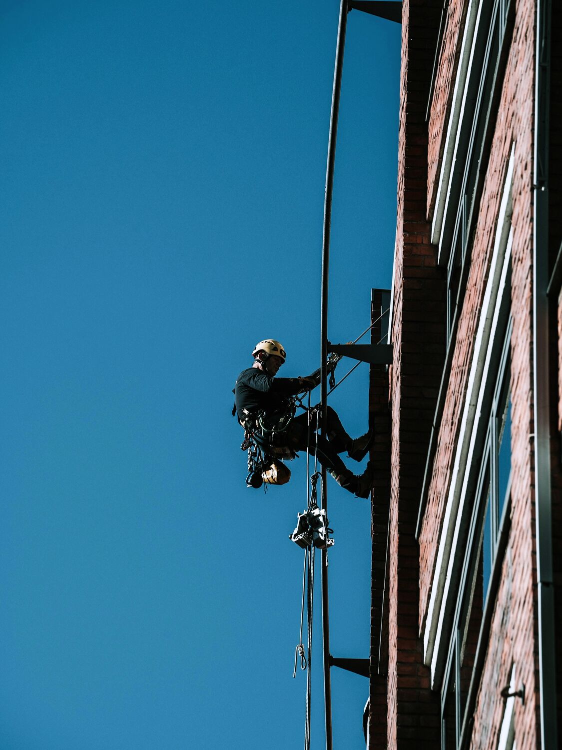A rope access technician abseiling down the face of a multi-storey building against a clear sky.