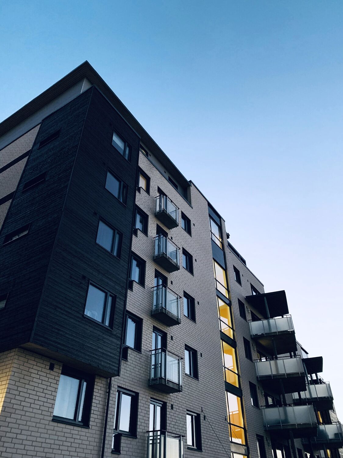 A modern low-rise apartment building with balconies, seen from below against blue sky.